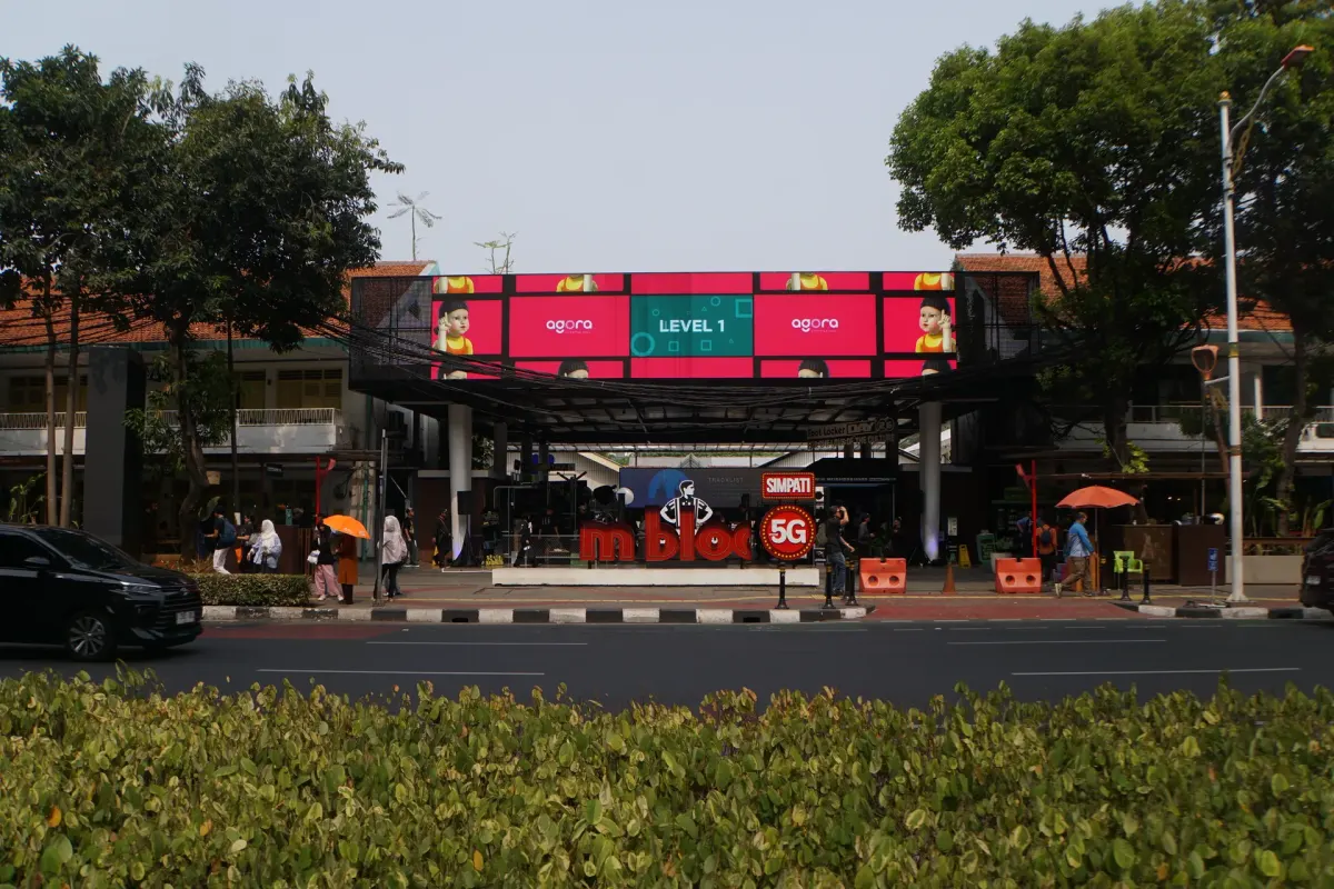 Layar LED memanjang di atas pintu masuk kawasan M Bloc, Jl. Panglima Polim, Jakarta Selatan, dengan pejalan kaki di trotoar