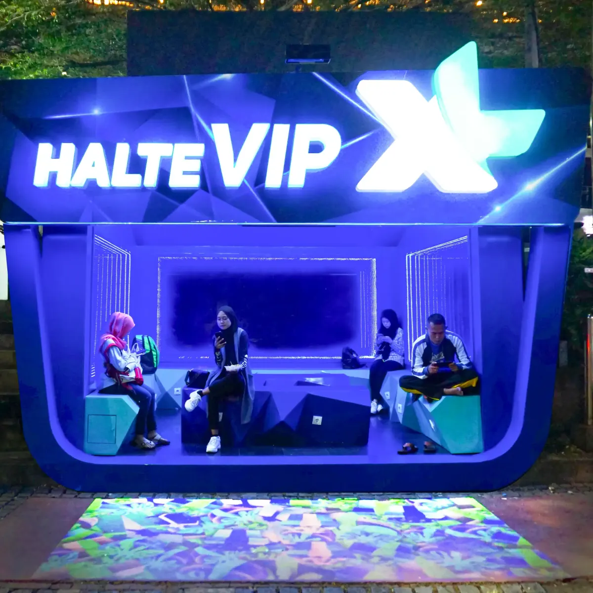 XL VIP bus shelter lit in blue at night, with a colourful image projected onto the pavement in front of it