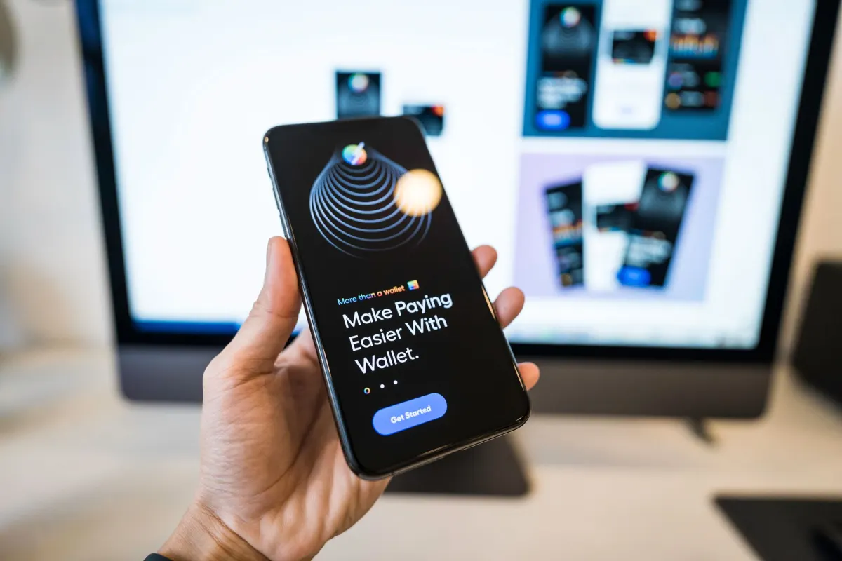 Hand holding a smartphone showing a digital wallet app onboarding screen, with a blurred monitor in the background