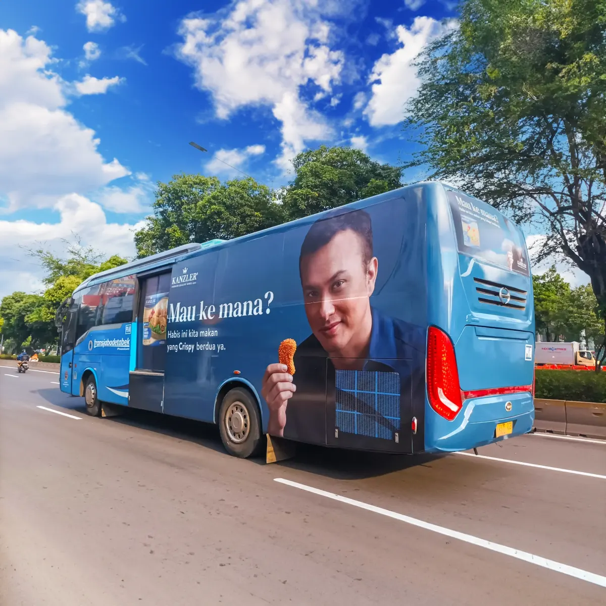 Blue Transjabodetabek Premium bus with a full-body ad wrap on its side and rear, driving on a main road under a clear sky