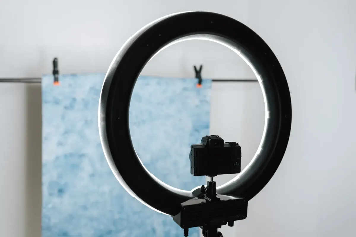 Kamera di atas tripod di tengah ring light dengan latar kain biru yang dijepit di depan dinding putih