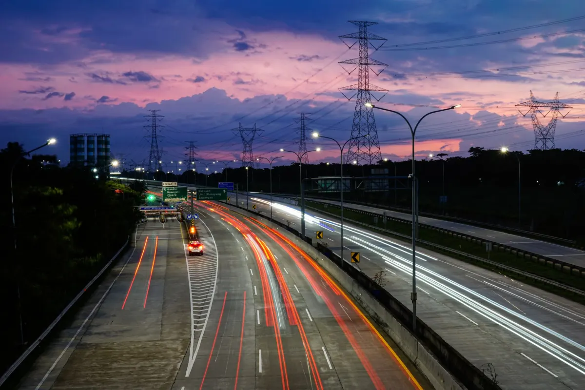 Toll road at dusk with vehicle light trails and power pylons under a purple sky
