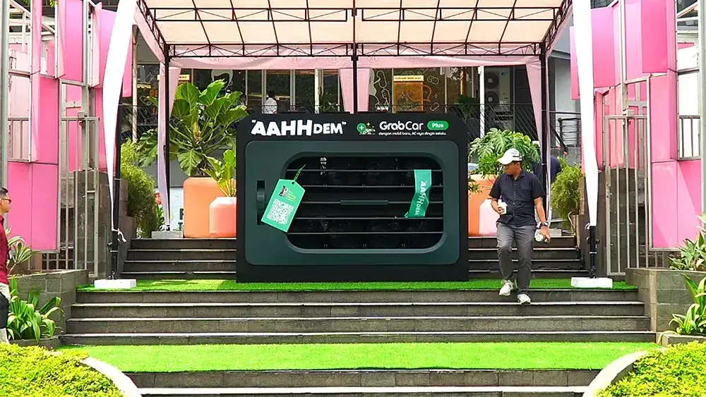 Grab on-ground creative branding: an oversized dark green air-conditioner unit on open plaza steps, framed by a white canopy and pink panels