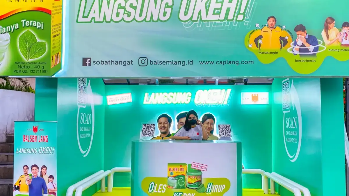 Halte bus yang didekorasi bertema produk balsem herbal dengan meja konter sampling di tengah, kode QR di dinding samping, dan roll banner di trotoar